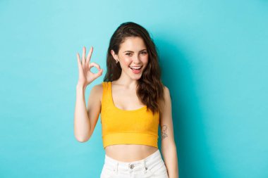 Kendine güvenen, gülümseyen, evet diyen, onay işareti gösteren, iyi bir seçim yapan, mavi arka planda duran genç bir kadının portresi.