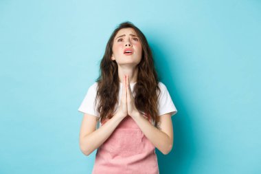 Umutsuz, genç, esmer bir kadın Tanrı 'ya yalvarıyor, dua ediyor ve umut dolu bir yüzle gökyüzüne bakıyor, rüya için yalvarıyor, mavi arka planda duruyor.