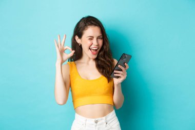 Teknoloji ve yaşam tarzı konsepti. Neşeli esmer kadın model evet diyor, göz kırpıyor ve işaret veriyor, cep telefonunu tutuyor, mavi arka planda duruyor.