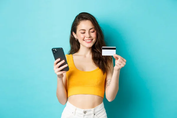 Chica con estilo con tarjeta de crédito de plástico sonriendo, pagando por el pedido en línea, compras en el teléfono inteligente, de pie sobre fondo azul — Foto de Stock