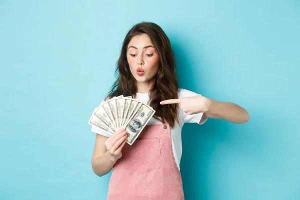 Emocionado linda chica sosteniendo el dinero y apuntando a los billetes de dólar, ir de compras, de pie sobre fondo azul — Foto de Stock