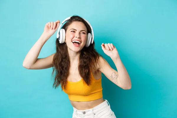 Mujer joven en ropa de verano escuchando música, usando auriculares y cantando junto a la canción favorita, bailando en auriculares, de pie sobre fondo azul — Foto de Stock