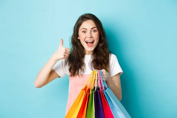 Εικόνα της χαρούμενης νεαρής γκλαμουράτης γυναίκας κατάστημα σε καταστήματα με εκπτώσεις, δείχνοντας τον αντίχειρα επάνω, κρατώντας τσάντες αγορών, κοιτάζοντας ενθουσιασμένος στην κάμερα, μπλε φόντο — Φωτογραφία Αρχείου