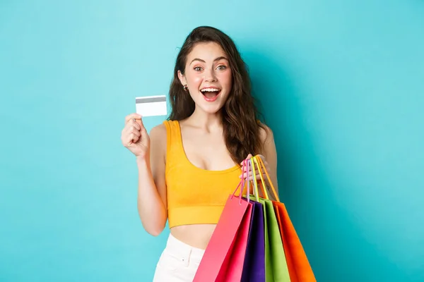 Young excited woman holding shopping bags and showing plastic credit card, buying things during promo deals, standing over blue background