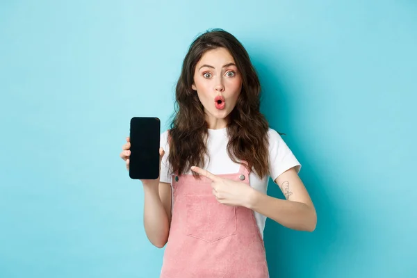 给人印象深刻的年轻现代女性指着智能手机屏幕，惊讶地看着相机，展示令人敬畏的应用程序，新的网上商店，站在蓝色的背景上 — 图库照片