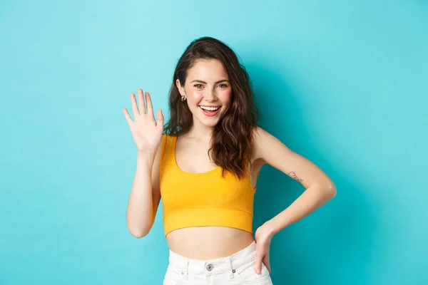 Summer holidays and emotions concept. Cheerful friendly woman with perfect body, waving hand and saying hello, making hi gesture, greeting you, standing over blue background