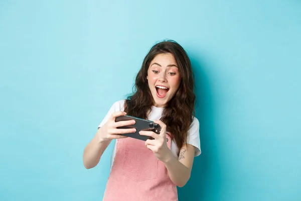 Retrato de uma jovem excitada jogando videogame móvel com as duas mãos, sorrindo divertido, olhando para a tela, em pé sobre o fundo azul — Fotografia de Stock