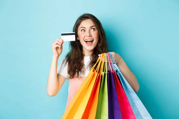 Glückliches brünettes Mädchen, Shopaholic zeigt ihre Plastikkreditkarte und Einkaufstüten, schaut verträumt auf Logo oder Banner des Geschäfts, steht über blauem Hintergrund — Stockfoto