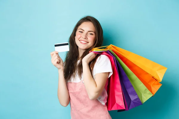 Jovem mulher satisfeita sorrindo, mostrando cartão de crédito de plástico e segurando sacos de compras, comprando com pagamento sem contato, de pé sobre fundo azul — Fotografia de Stock