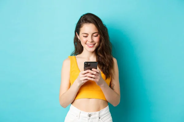 Mulher moderna atraente conversando no telefone móvel, olhando para a tela com sorriso concurso, mensagens no namoro aplicativo, de pé sobre fundo azul — Fotografia de Stock