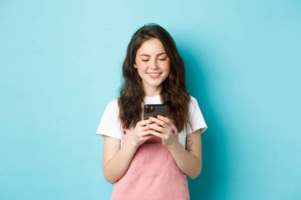 Portrét mladé ženy pomocí smartphonu, čtení zprávy nebo chatování v aplikaci, stojící nad modrým pozadím a šťastný úsměv — Stock fotografie