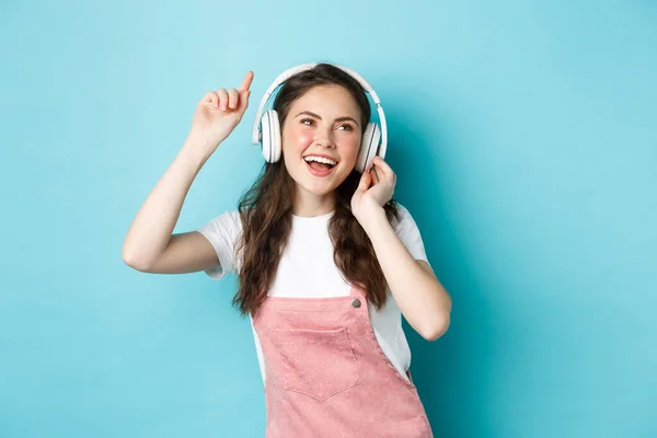 Mujer joven bailando y escuchando música en auriculares, levante la mano y sonriendo despreocupado, disfrutando de la canción favorita, de pie sobre fondo azul — Foto de Stock