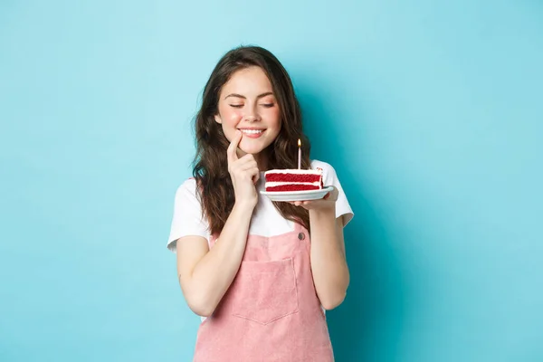 Fiestas y celebración. Chica de cumpleaños sonriente celebrando, mirando la torta de ensueño con vela, pidiendo deseo, quiere morder el postre, de pie sobre el fondo azul —  Fotos de Stock