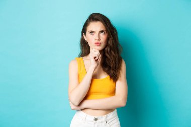 Yazlık kıyafetli çekici bir kızın portresi düşünme, karar verme, kaşlarını çatma ve sol üst köşeye dalgın bir şekilde bakma, mavi arka planda dikilme.