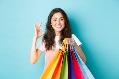 İndirimli mağazalardan alışveriş yapan mutlu, genç bir kadının resmi. İyi işaretler gösteriyor, alışveriş torbaları tutuyor, kameraya heyecanlı bakıyor, mavi arka plan