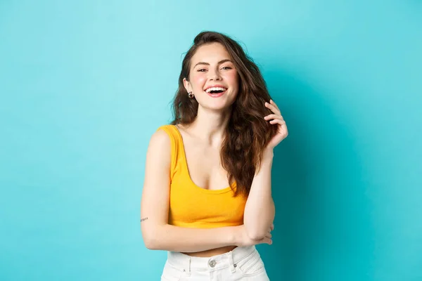 Concepto de verano y estilo de vida. Retrato de mujer atractiva riendo y jugando con su pelo, de pie en la parte superior recortada de color amarillo, ir de vacaciones, de pie sobre fondo azul —  Fotos de Stock