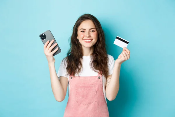 Sonriendo linda chica que muestra la tarjeta de crédito de plástico y el teléfono inteligente, pedir algo en la tienda a través de la aplicación móvil, tienda en línea, o pagar por el pedido, de pie contra el fondo azul — Foto de Stock