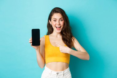 Şuna bir bak. Gülümseyen genç bir kadın boş akıllı telefon ekranını işaret ediyor, internetten reklam öneriyor, mavi arka planda duruyor.