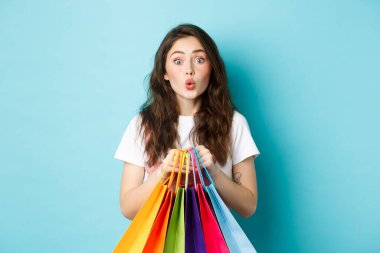 Heyecanlı genç kadın portresi mağazada iyi bir indirim teklifi görüyor ve kameraya bakıyor, alışveriş torbaları tutuyor, mavi arka planda duruyor.