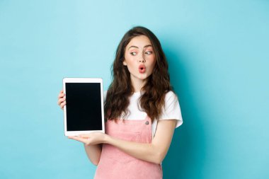İlgi çekici kız dijital tablet ekranı izlerken size gösteriyor, online tanıtım teklifiyle eğleniyor, ekranda bir şey gösteriyor, mavi arkaplan