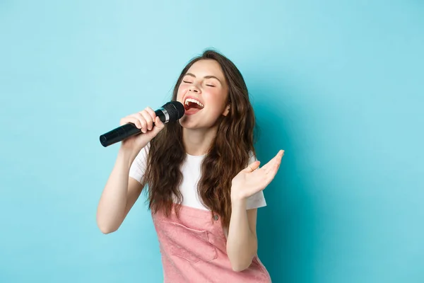 Mujer hermosa despreocupada realizar canción, cantando en el micrófono con pasión, jugando karaoke, de pie sobre fondo azul — Foto de Stock