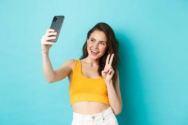 Conceito de tecnologia e estilo de vida. Elegante jovem mulher tomando selfie em suas roupas de verão, mostrando v-sinal na câmera do smartphone, de pé sobre fundo azul — Fotografia de Stock