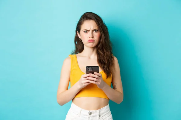 Confused and freak out young woman frowning, grimacing after reading strange message on smartphone, standing over blue background