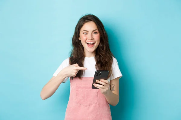 Retrato de bonito e alegre menina apontando o dedo para o telefone, usando aplicativo de compras de smartphones, de pé sobre fundo azul — Fotografia de Stock