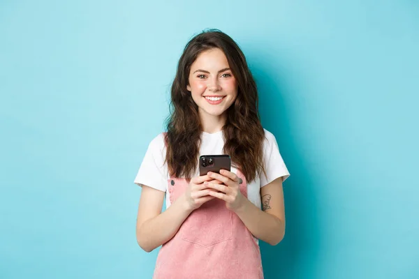 Mladá roztomilá dívka drží mobilní telefon, usmívá se a dívá se do kamery. Žena pomocí aplikace smartphone, chatování na sociálních sítích nebo nakupování online, modré pozadí — Stock fotografie