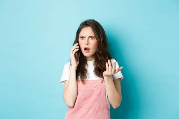 Ritratto di donna confusa e scioccata ricevere cattive notizie mentre si parla su smartphone, ricevere cattive telefonate, in piedi su sfondo blu — Foto Stock