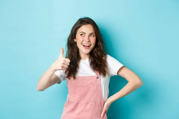 Sorrindo menina adolescente bonito com blush bonito e glamour compõem, mostrando polegar para cima na aprovação, olhar para o banner canto superior esquerdo, recomendando loja, de pé contra fundo azul — Fotografia de Stock