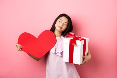 Sevgililer Günü konsepti. Romantik ve hassas Asyalı genç kız gülümsüyor, gözlerini kapatıyor ve sevgilisinin hediyeleriyle hayaller kuruyor, hediyesini tutuyor ve büyük kırmızı kalp kesimi, pembe arka plan
