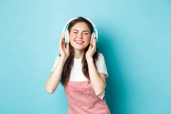 Cute kaukaskie dziewczyna w strój wiosna, słuchanie muzyki w słuchawki, uśmiechając zadowolony z aparatu, stojąc nad niebieskim tle — Zdjęcie stockowe