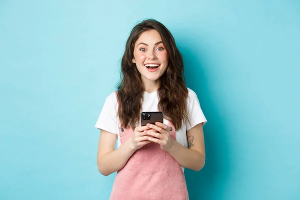 Retrato de menina morena animado olhando maravilhado para a câmera ao usar o smartphone, segurando telefone celular e sorrindo alegre, de pé contra o fundo azul — Fotografia de Stock