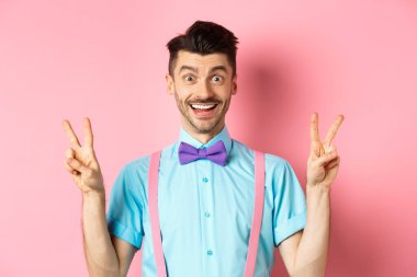 Bıyıklı ve papyonlu neşeli genç adam barış veya zafer jestleri gösteriyor, kameraya mutlu bir şekilde gülümsüyor, pembe arka planda duruyor.