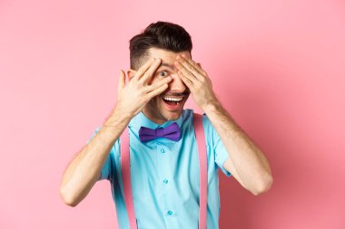 İlgi çekici papyonlu komik adam, parmaklarının arasından ilginç bir şeye bakıyor, havalı bir şeye bakıyor, pembe arka planda duruyor.