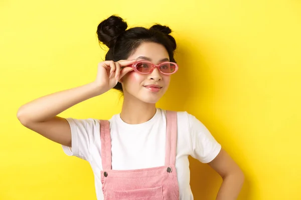 Όμορφο κορίτσι από την Ασία με κομψά γυαλιά ηλίου και χαμόγελο, φορώντας μοντέρνα καλοκαιρινά ρούχα, στέκεται σε κίτρινο φόντο — Φωτογραφία Αρχείου