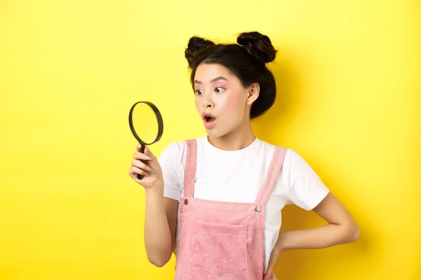 Opgewonden aziatische vrouw met heldere glamour make-up, kijk door vergrootglas verbaasd, staren opzij, staan op gele achtergrond — Stockfoto