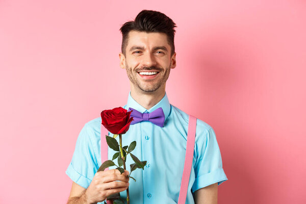 Привлекательный модный парень ждет свидания на свидании влюбленных, держит красную розу и улыбается, стоя на романтическом розовом фоне. День святого Валентина
