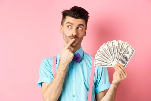 Hombre pensando en ir de compras, mirando pensativo mientras sostiene el dinero en dólares, de pie sobre el fondo rosa — Foto de Stock