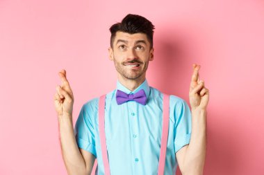 Papyonlu gülümseyen genç adam, dua ediyor, dua ediyor, bakıyor, şans diliyor, pembe arka planda umutla bekliyor.