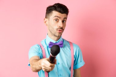 Yakışıklı erkek sunucu mikrofonla elini uzatıyor, röportaj ya da yorum istiyor, pembe arka planda duruyor.