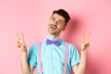 Bıyıklı ve papyonlu, barış ya da zafer işaretleri gösteren ve pembe arka planda poz veren mutlu sevimli adam.