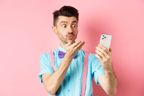기술의 개념. 비디오 채팅으로 에어 키스를 보내고 스마트폰 카메라에서 몸을 날려 분홍색 배경에 서 있는 웃긴 남자 — 스톡 사진