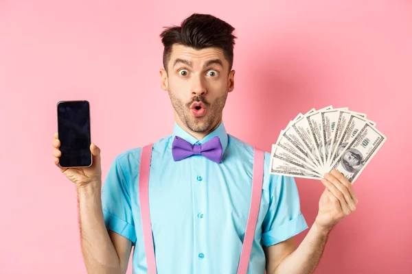 Concepto de comercio electrónico y compras. Hombre sorprendido mostrando pantalla de teléfono inteligente en blanco y dinero, diciendo wow con la cara sorprendida, echa un vistazo a la oferta en línea, fondo rosa — Foto de Stock