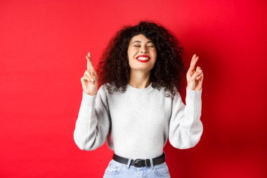 Kırmızı dudaklı, kıvırcık saçlı, umut dolu ve pozitif bir kadın, şans dileyen ve dilek tutan, rüya için dua eden, heyecanlı, kırmızı arka planda gülümseyen.