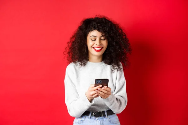 Chica positiva feliz charlando por teléfono, leyendo el mensaje y sonriendo, utilizando la aplicación de medios sociales, de pie sobre fondo rojo — Foto de Stock