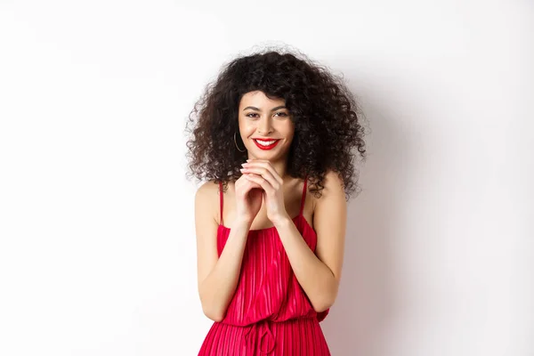 Mujer joven astuta con el pelo rizado, dedos de campanario y sonriente retorcido, con un plan, de pie en vestido rojo sobre fondo blanco — Foto de Stock