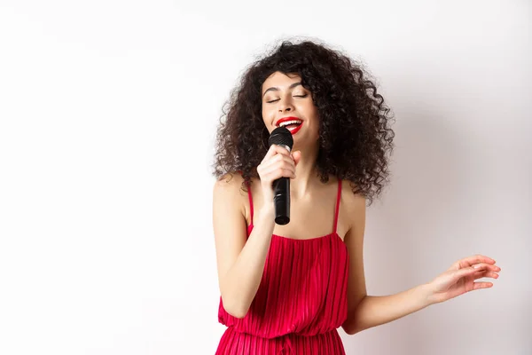 Felice donna elegante in abito rosso esibendosi con microfono, canto karaoke e sorridente, in piedi su sfondo bianco — Foto Stock