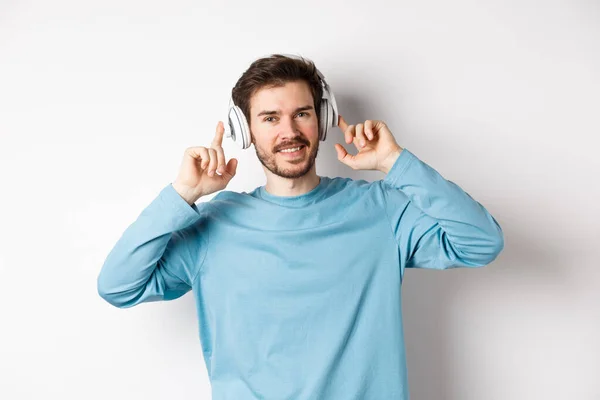 Homem moderno atraente com barba, sorrindo satisfeito, tocando fone de ouvido na cabeça enquanto ouve música, de pé sobre fundo branco — Fotografia de Stock